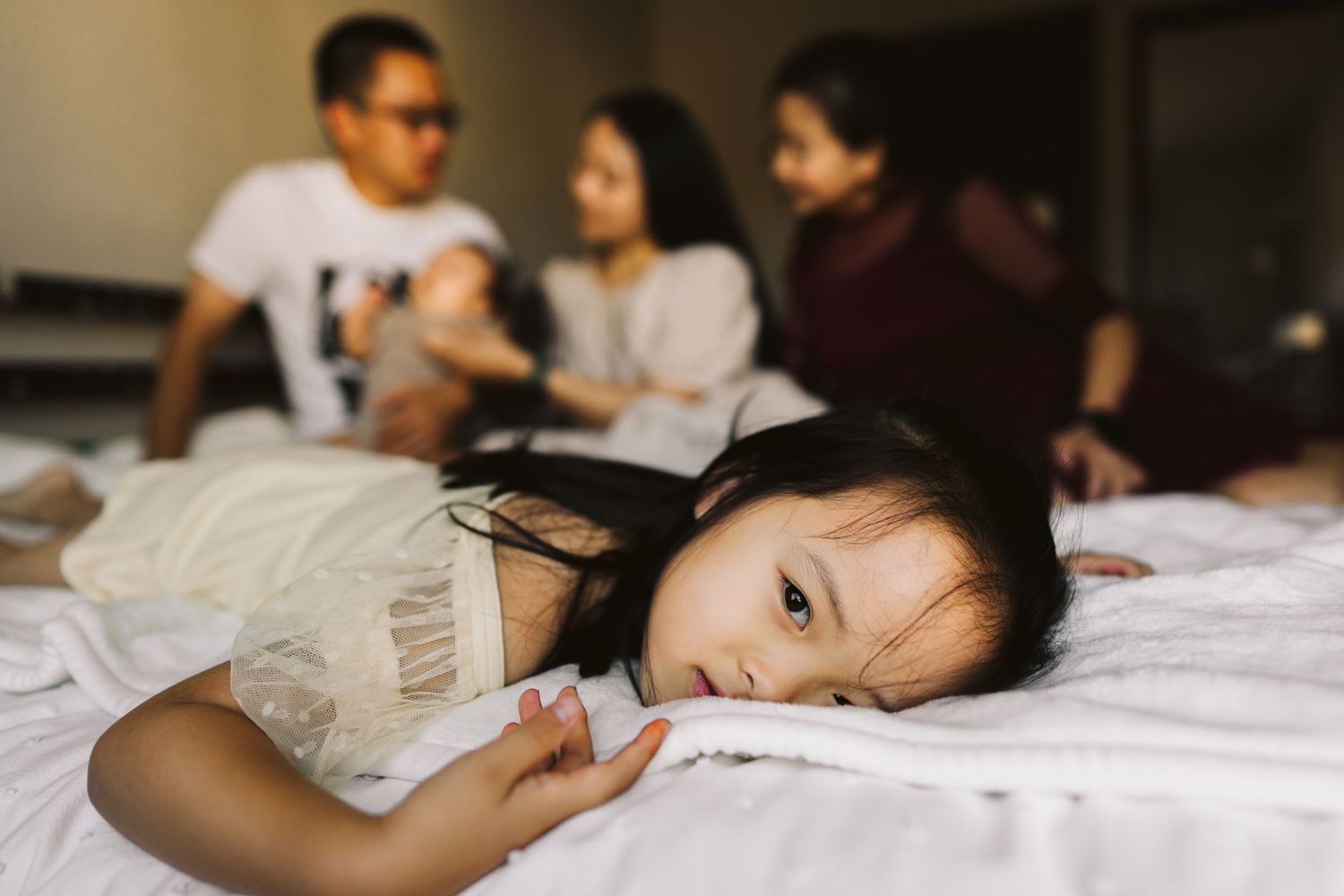A young child in white rests on a bed while adults gather and chat in the background.