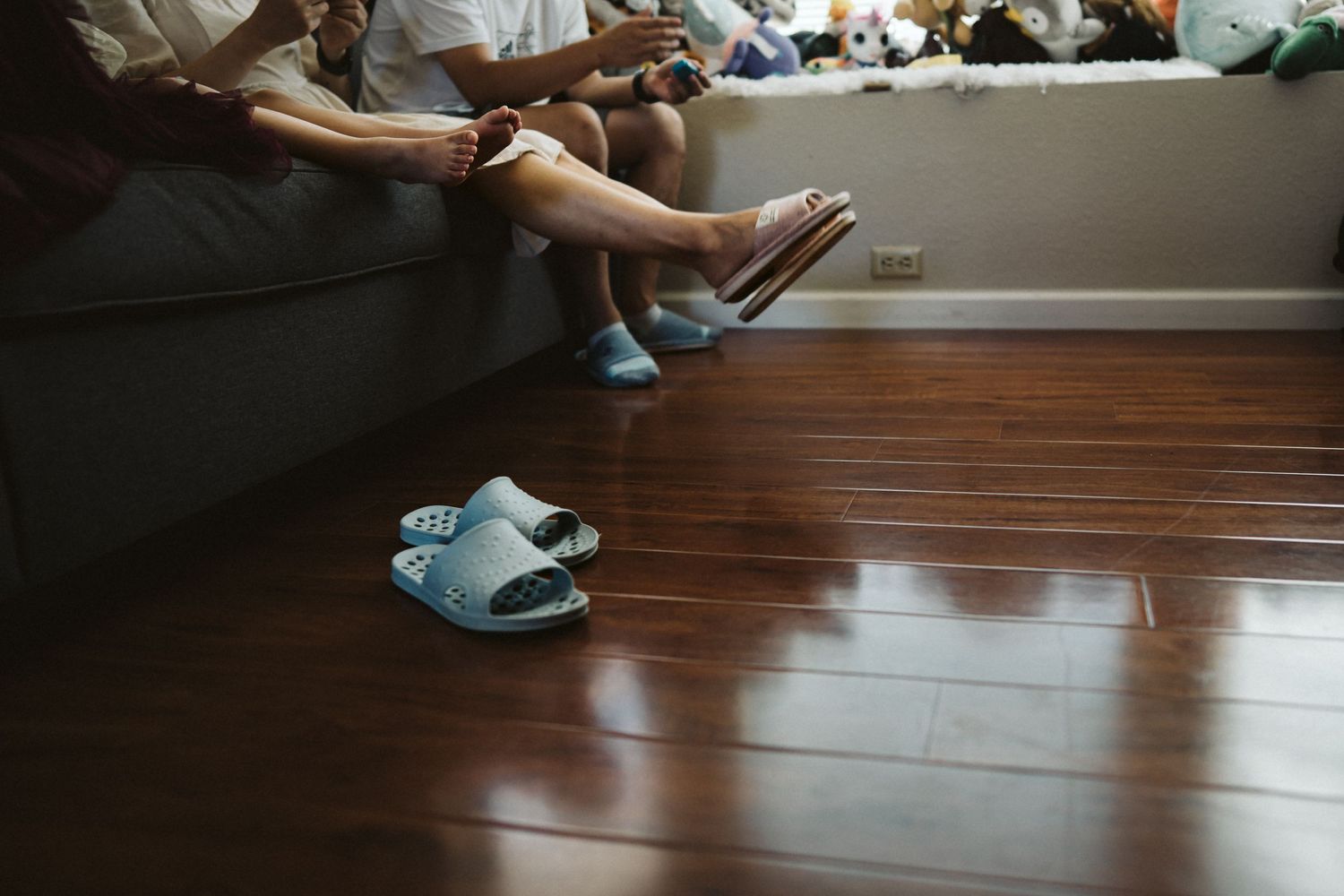 Bare feet resting on a dark couch with light blue slippers on hardwood floor in a living room.