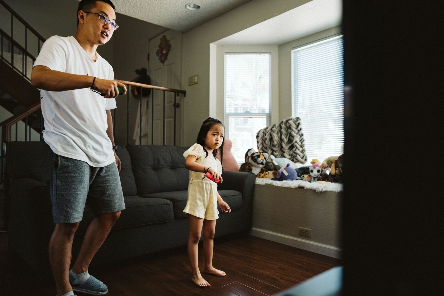 Two people stand playing with video game controllers in a living room near a bright window.