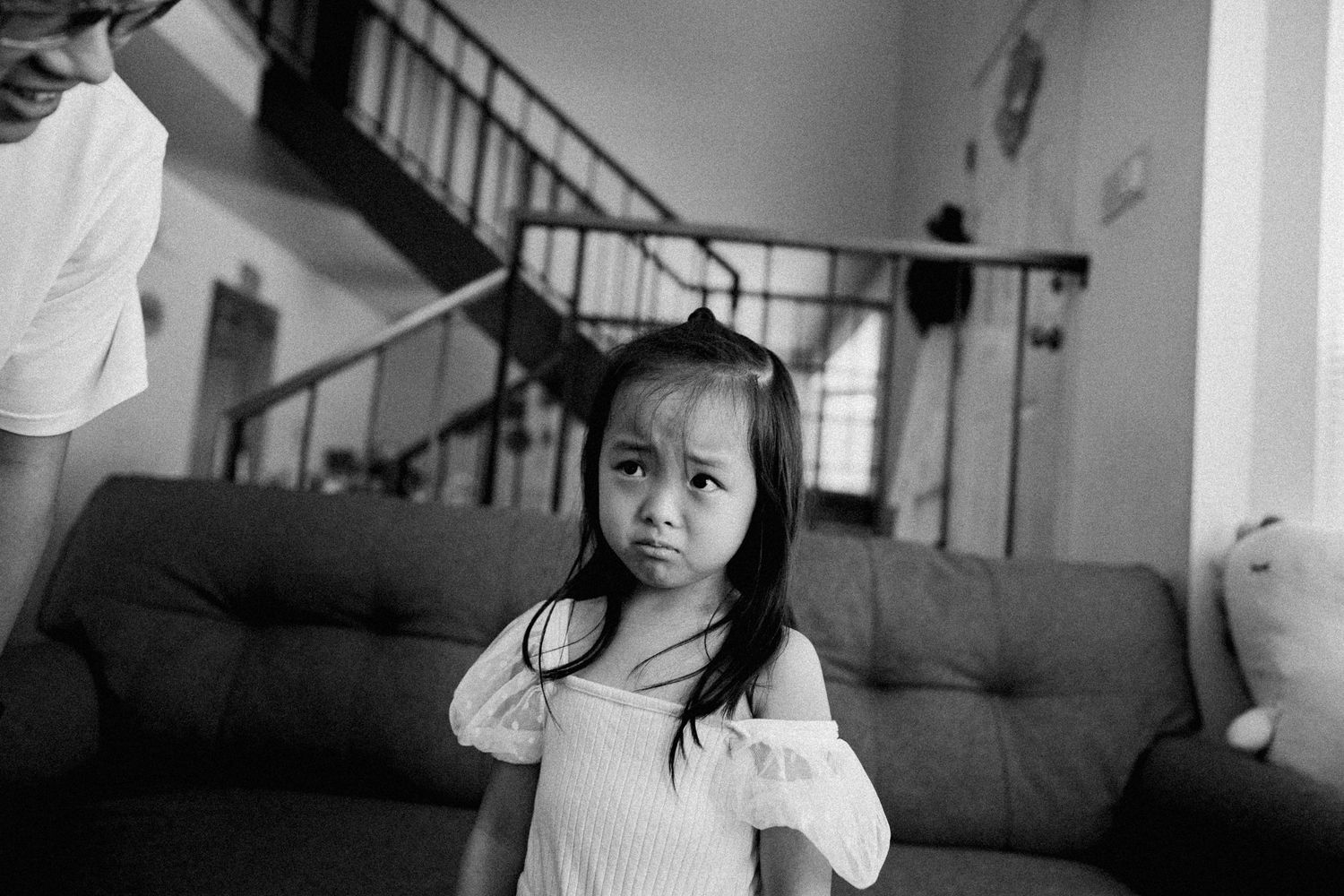 Young girl in a white off-shoulder dress standing in a living room with a dark couch and staircase.