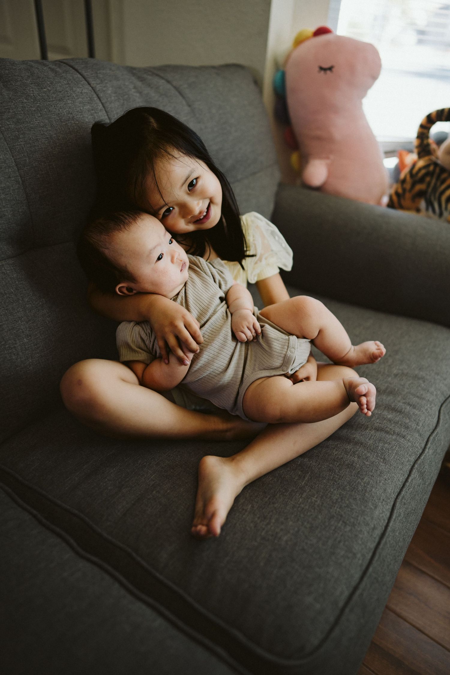 Two children sit together on a dark gray couch with stuffed animals visible in a cozy home setting.