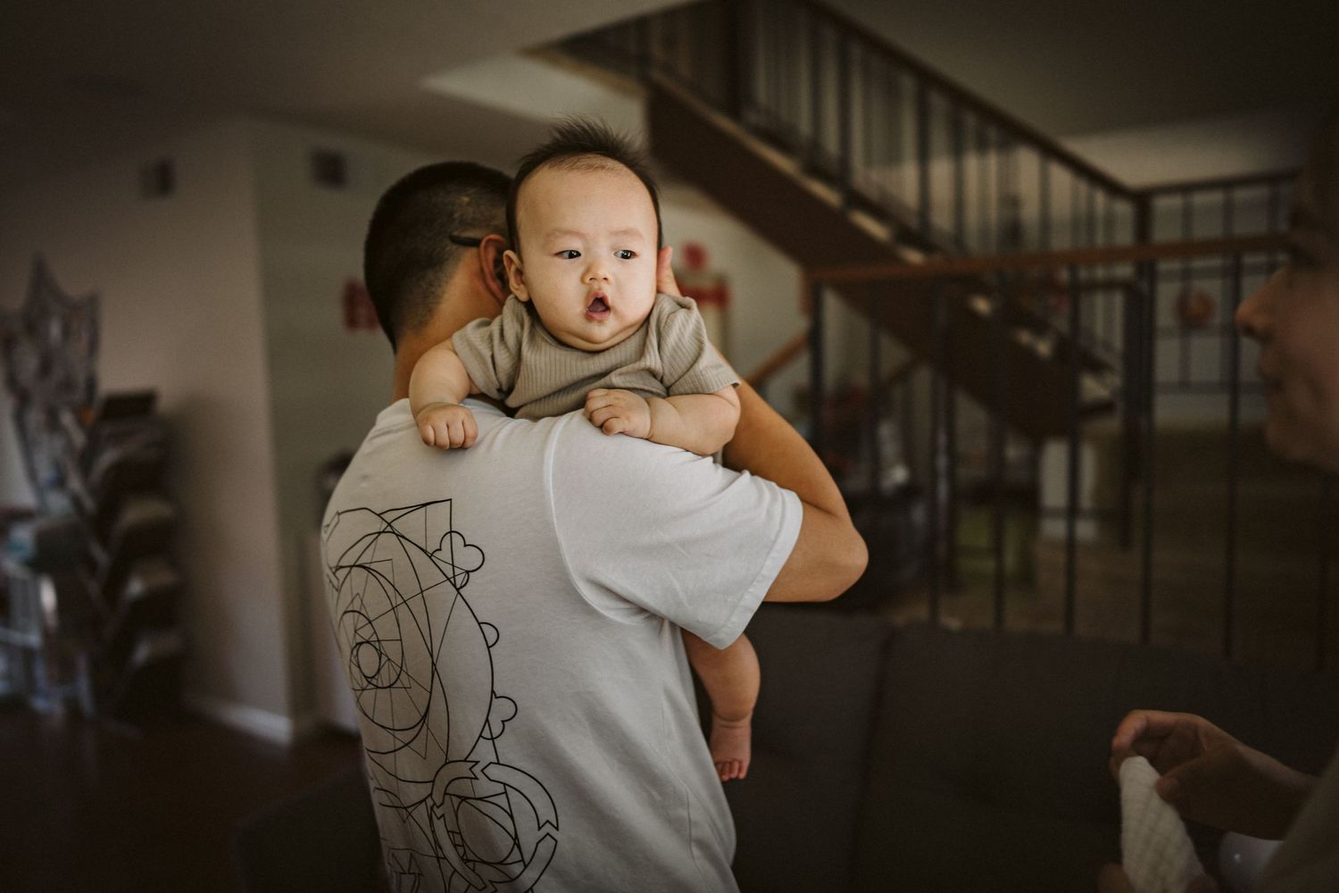 In a dimly lit home, someone holds a small baby over their shoulder wearing light colored clothing near a staircase.