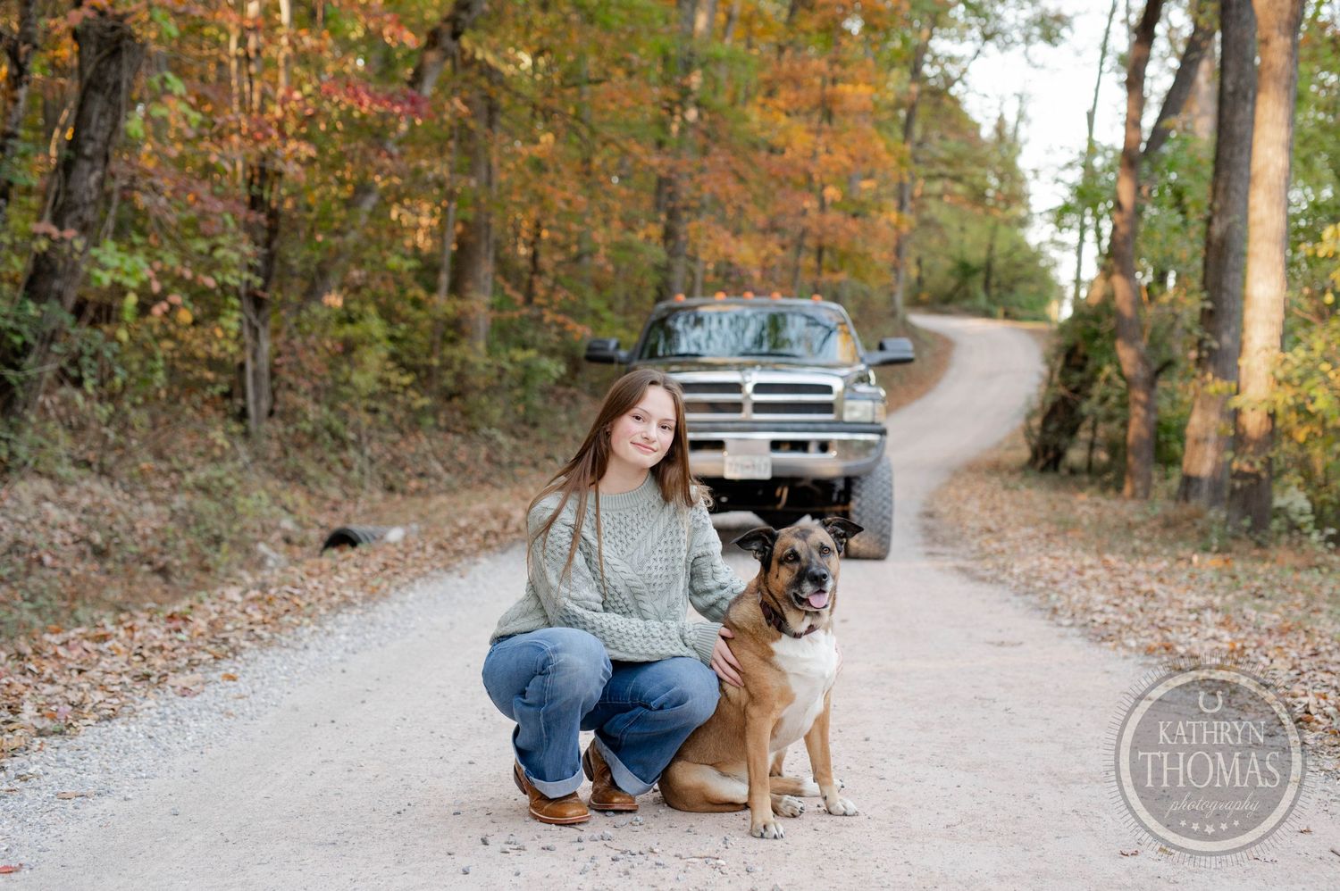 A person kneels beside a brown dog on a gravel road with a truck in the background.