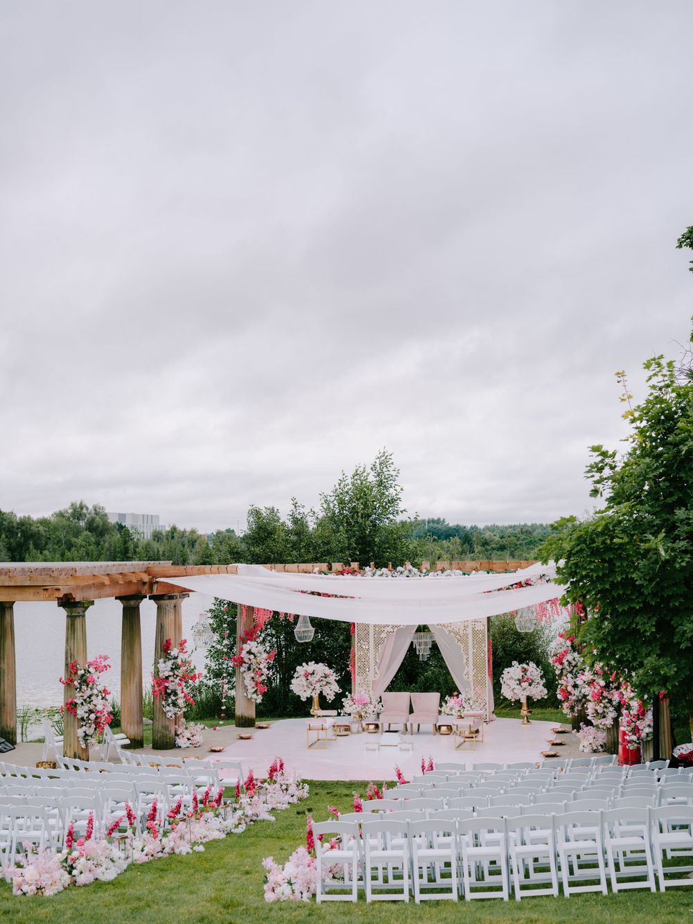 Elegant outdoor wedding ceremony setup with white canopy, wooden pergola and pink floral decorations on a cloudy day.