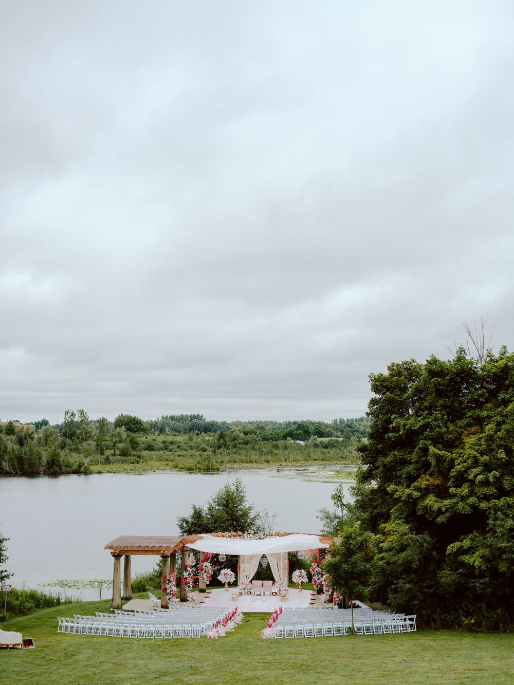 Outdoor wedding ceremony setup with a rustic wooden arch and white chairs overlooking a serene lake on a cloudy day.