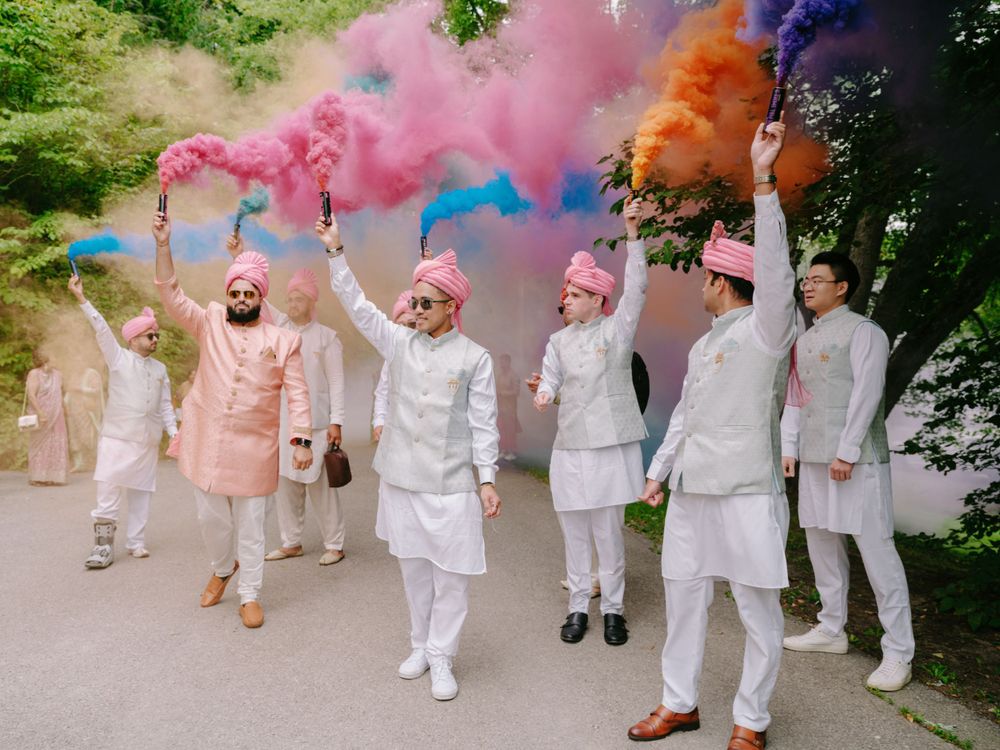 A group in traditional Indian attire walks along a path while holding colorful smoke bombs creating a vibrant display.