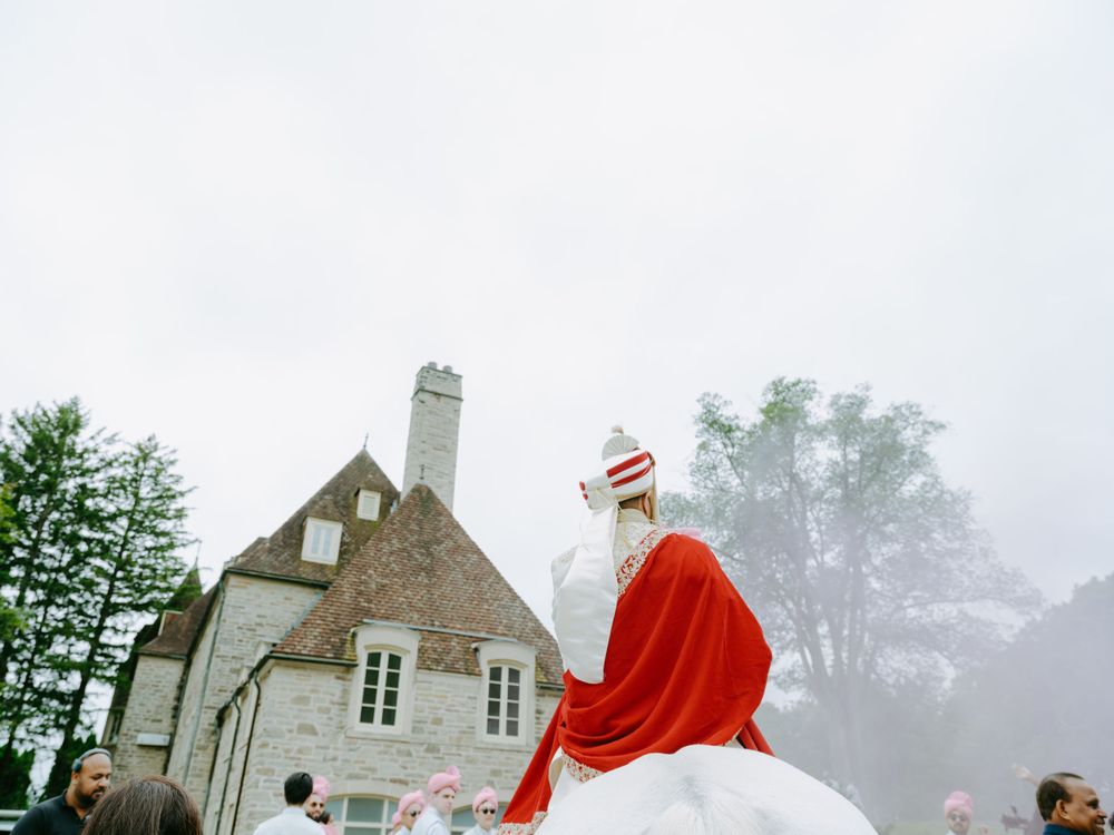 A figure in a red robe and mitre rides a white horse in front of a stone house on a misty day.