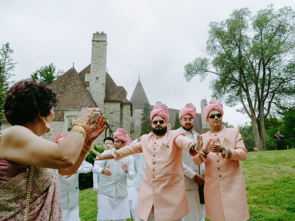 Wedding party wearing light pink and white outfits celebrates on a lawn with historic stone building in background.