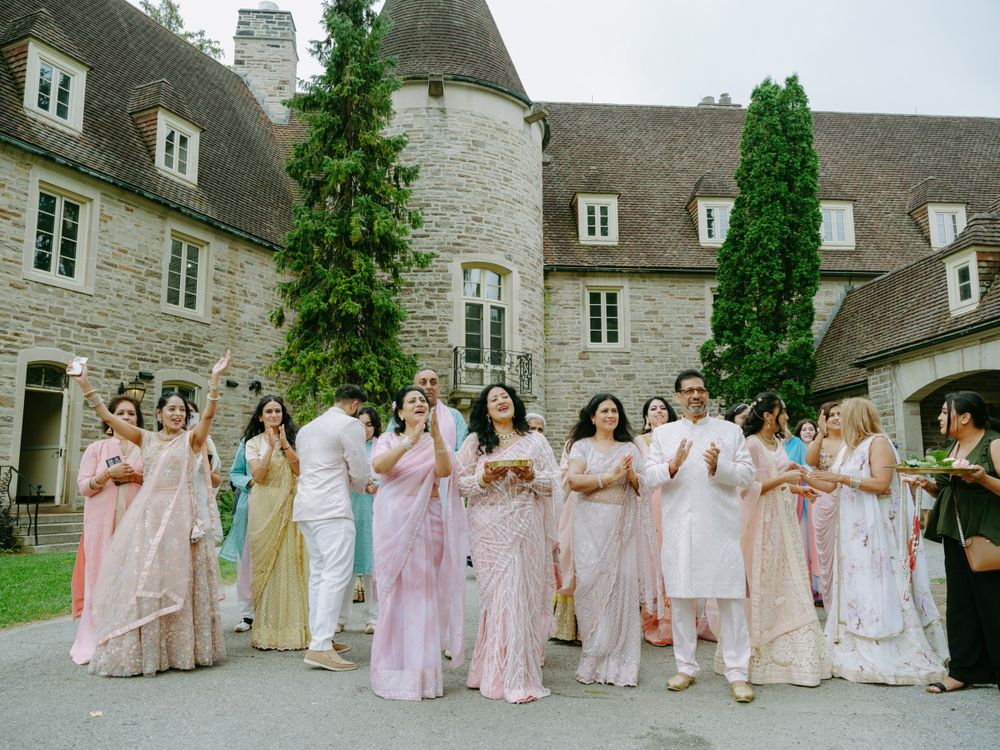 A row of bridesmaids in pastel pink and cream dresses stand outside a grand stone chateau with dormer windows.
