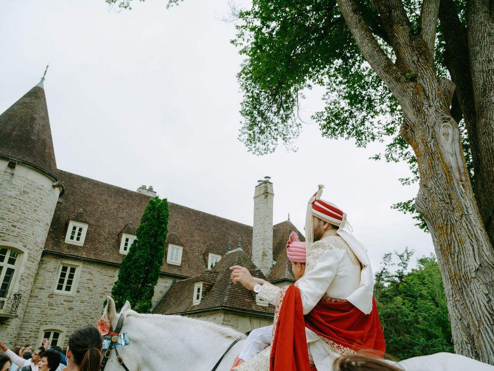 A figure in red and white traditional ceremonial attire rides a white horse in front of a medieval stone castle building.