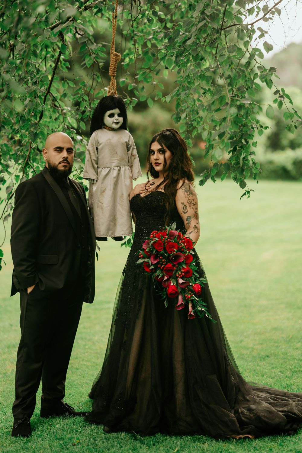 A dark gothic wedding photo featuring a black gown, red roses, and a creepy doll hanging from tree branches in nature.