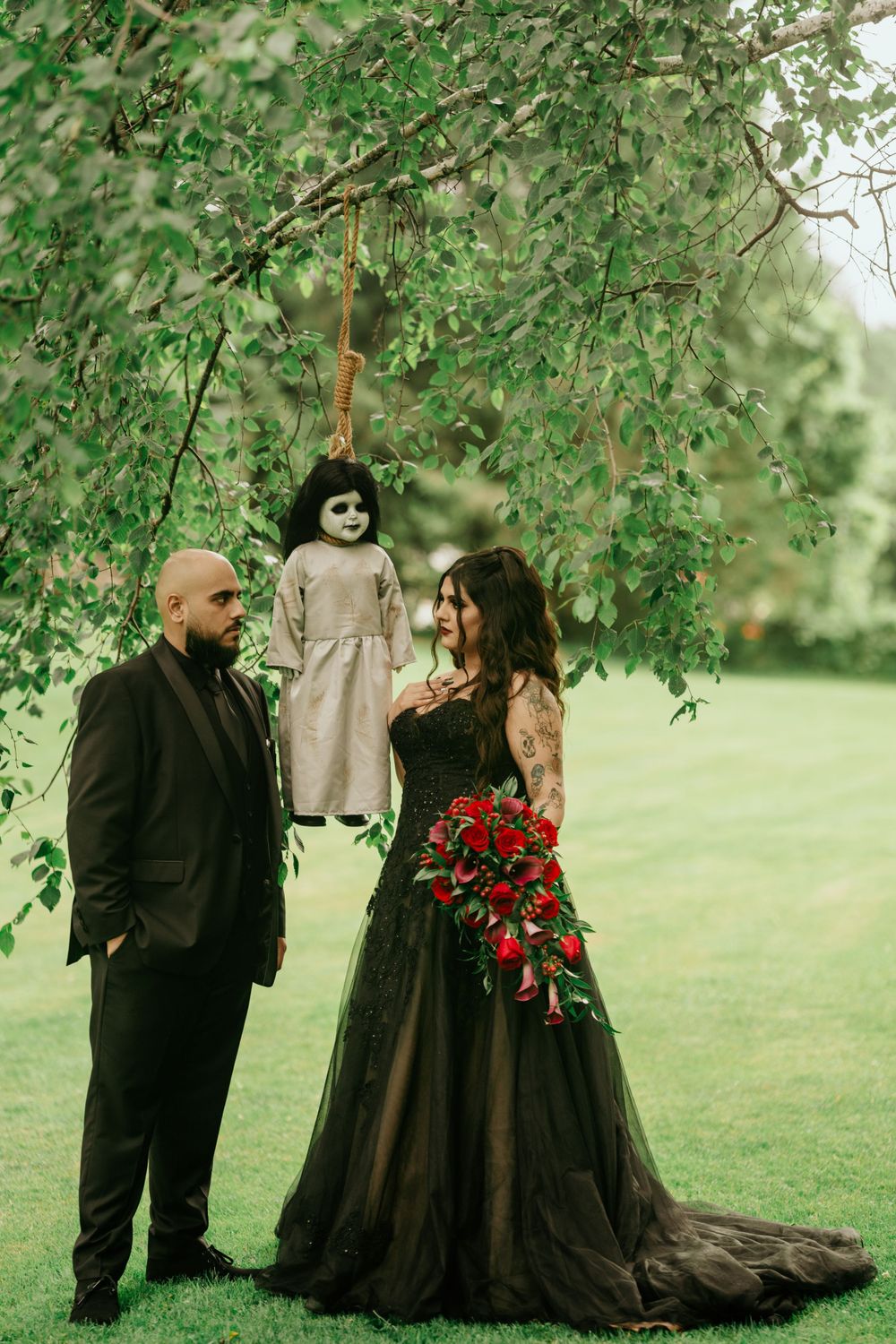 Gothic wedding couple poses under willow tree branches with hanging vintage doll and dramatic black dress.