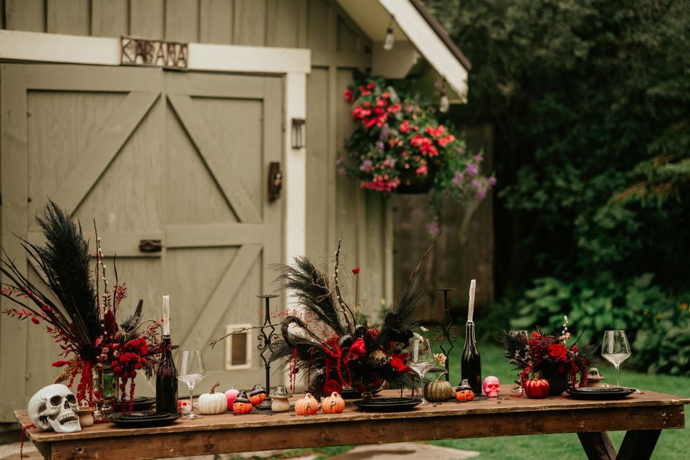 A rustic outdoor wooden table with black and red floral centerpieces against a barn door with hanging flower basket.