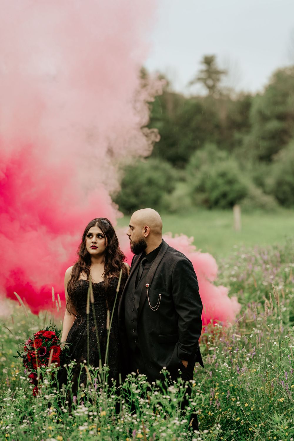 Dramatic wedding photoshoot with red smoke bombs in a wildflower field featuring gothic black attire.