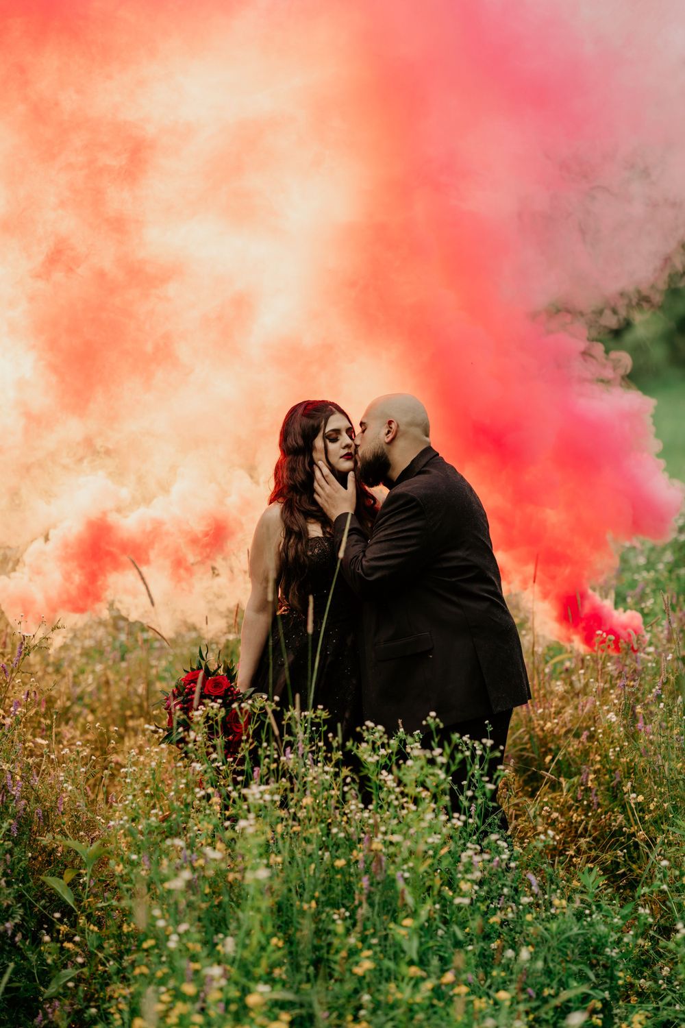 A romantic couple poses in a meadow with vibrant orange and pink smoke bombs creating a dramatic backdrop for their photo.