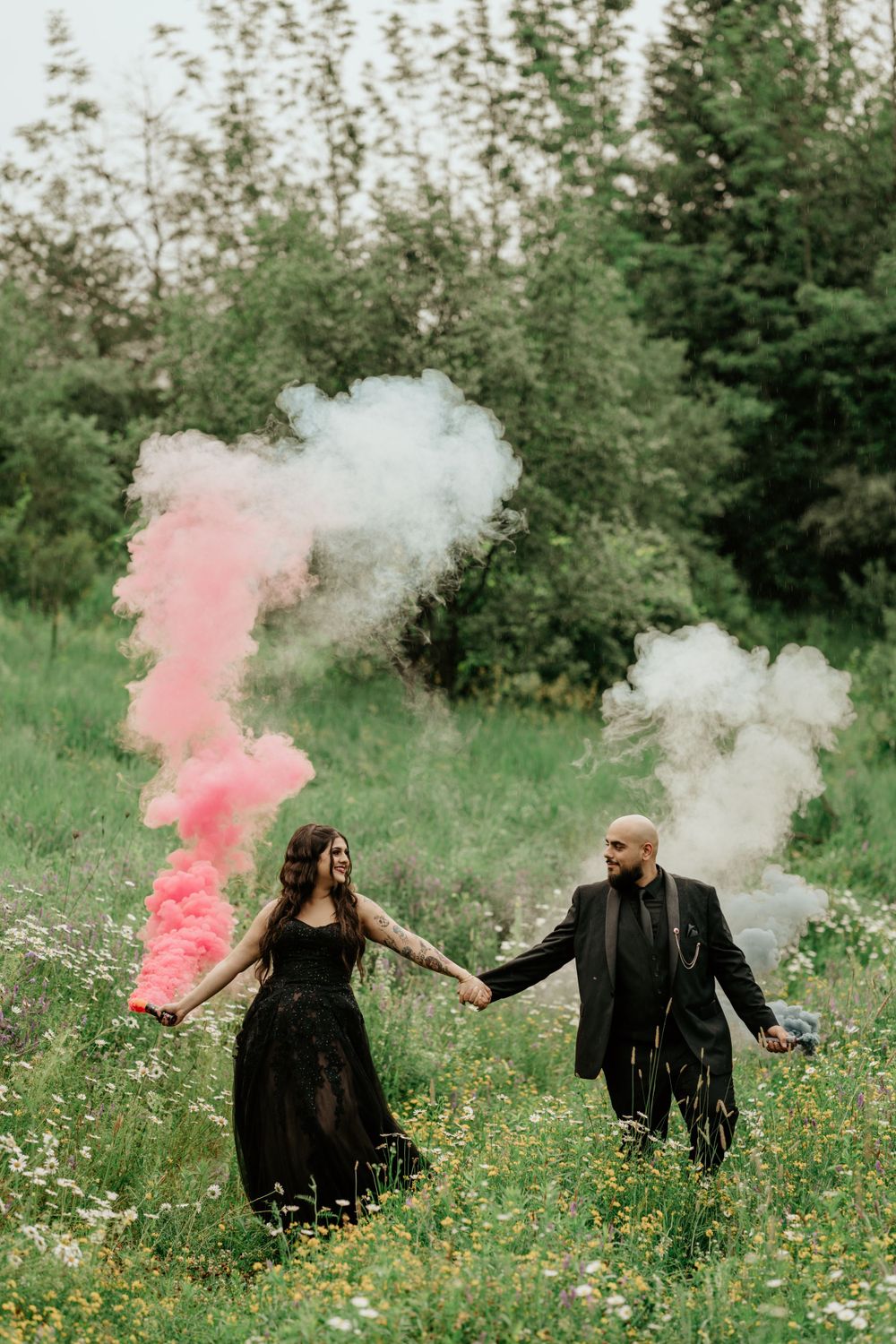 Couple in black gothic attire hold hands in a meadow with pink and white smoke bombs creating dramatic effect.