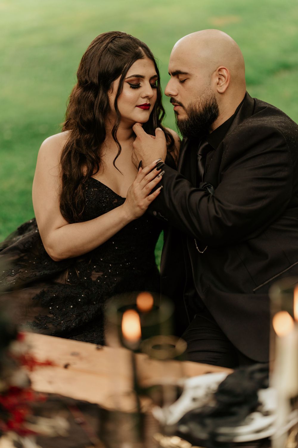 Romantic couple in elegant black attire share an intimate moment during outdoor photoshoot with candles and greenery.