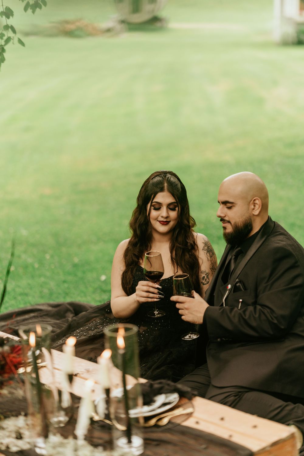 A couple in black attire shares drinks during an outdoor picnic with romantic decorations and candles.