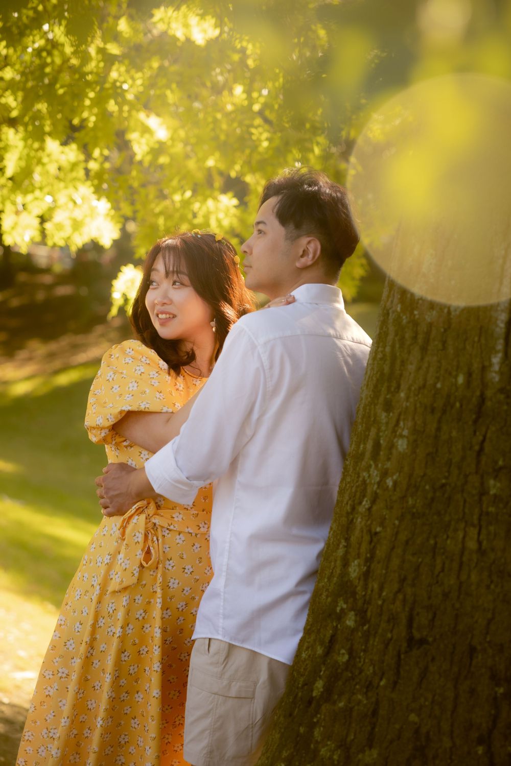 A romantic couple embraces beneath sunlit trees in a garden, with her wearing a yellow summer dress.