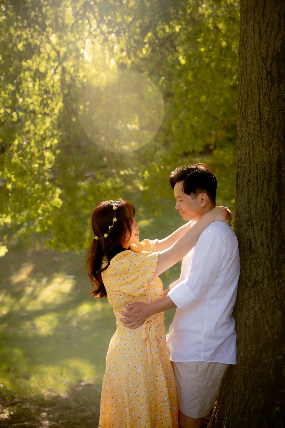 A tender moment captured between a couple against a tree trunk with golden sunlight filtering through leaves.