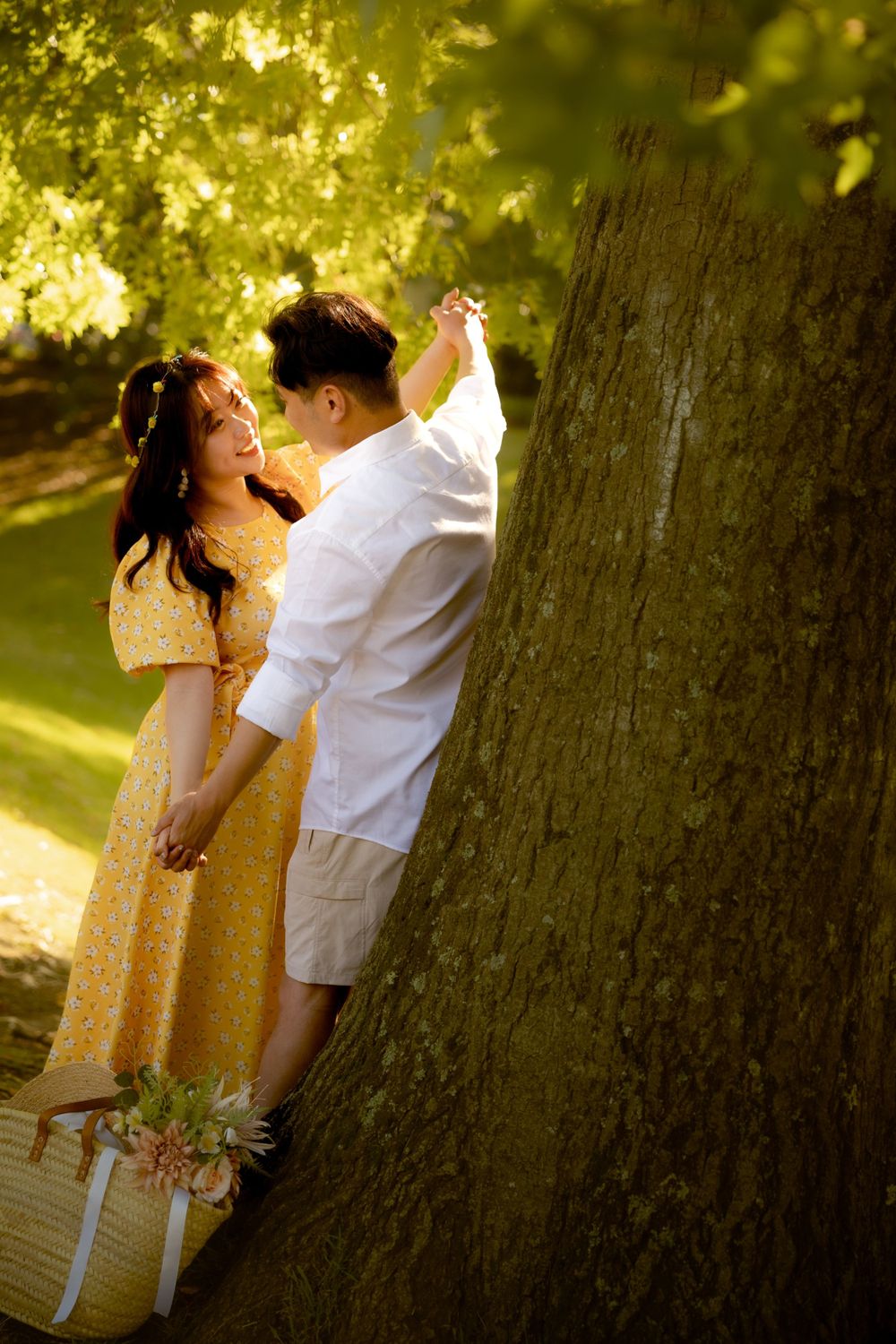 A couple in coordinated white and yellow outfits share a playful dance by a large tree.