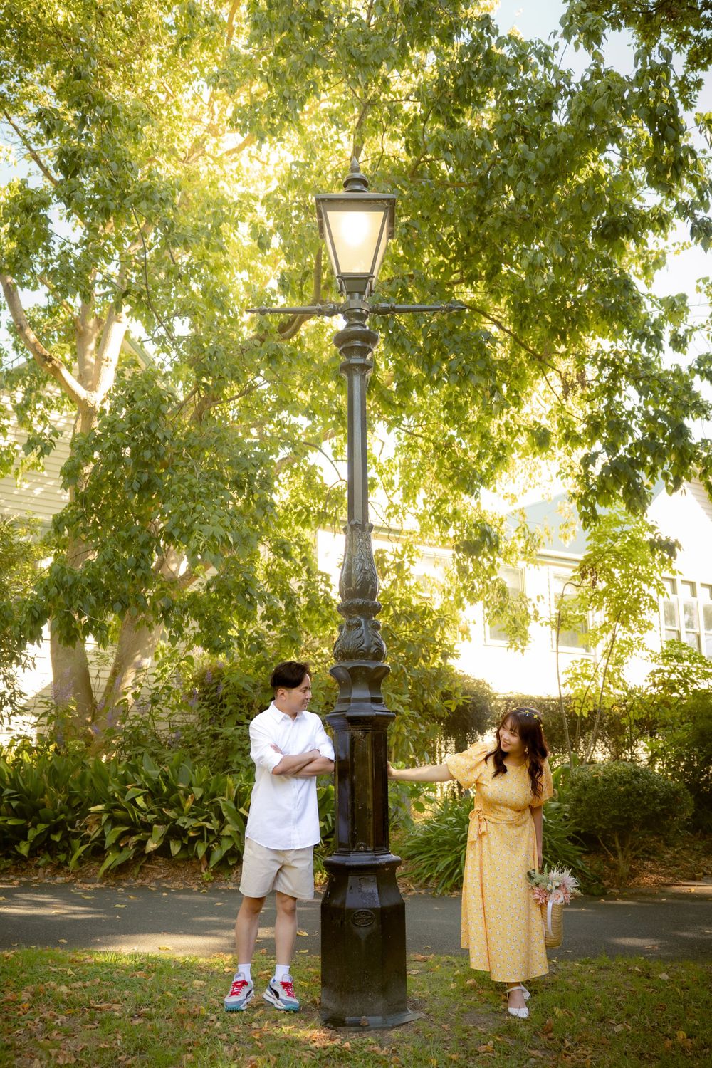 A couple stands on opposite sides of an ornate vintage lamp post in a leafy park setting.
