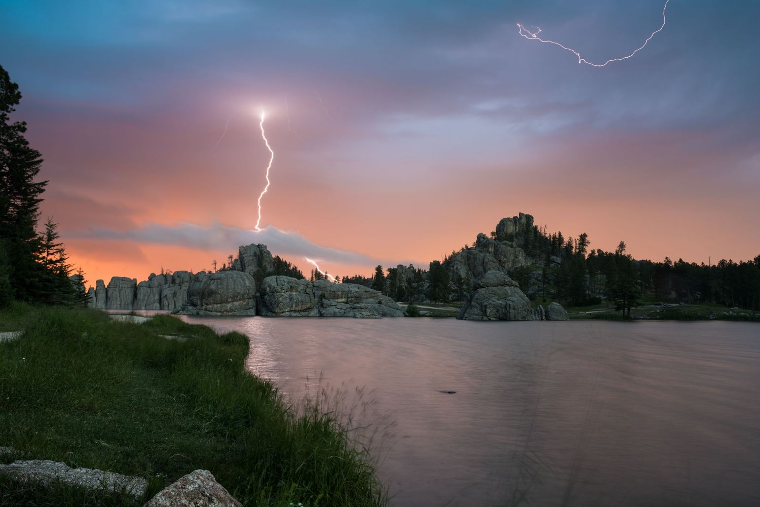 A dramatic lightning strike illuminates a lakeside landscape at sunset with rocky outcrops and silhouetted trees.