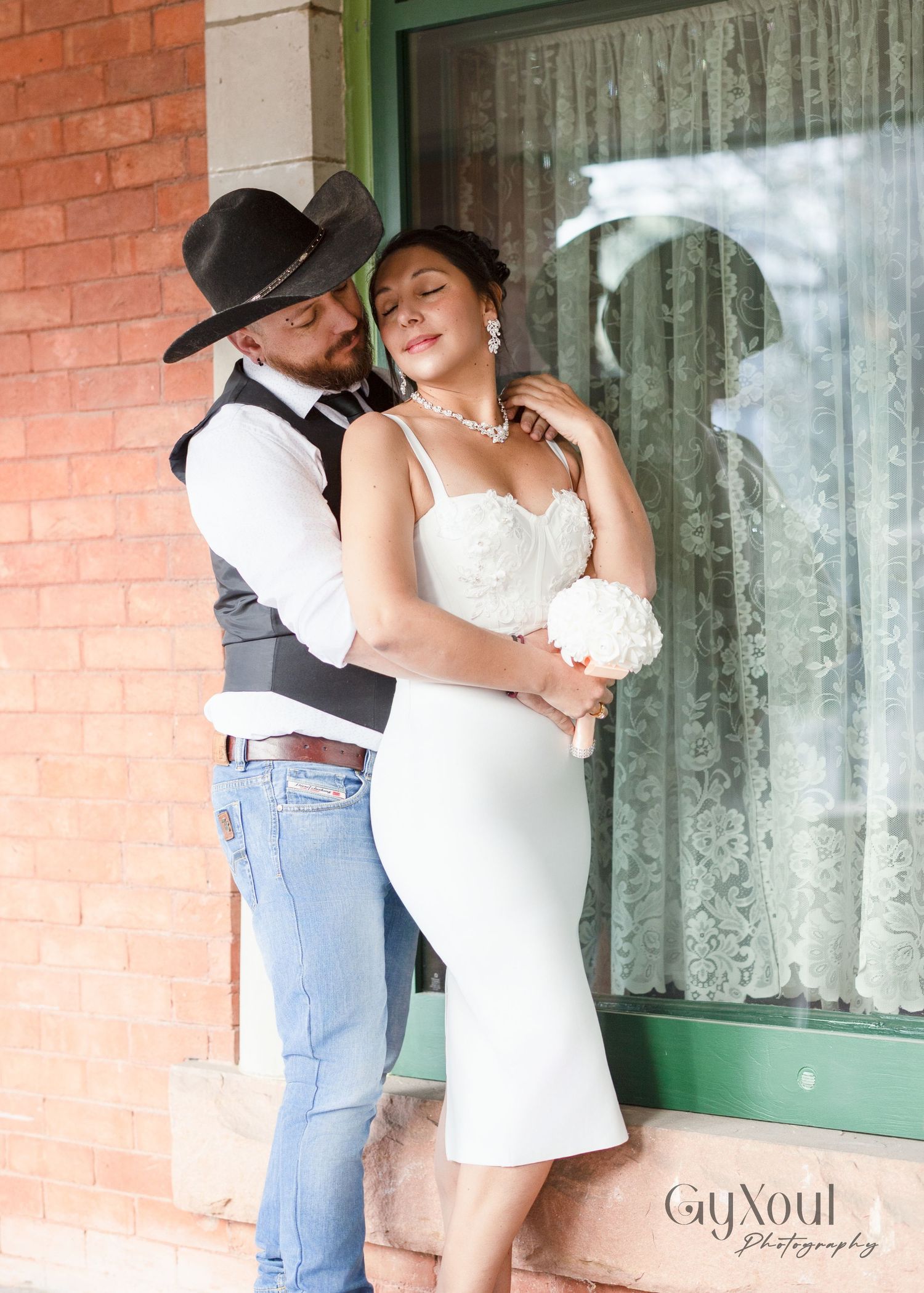 A romantic western wedding moment captured outside a brick building with lace curtains in the window.
