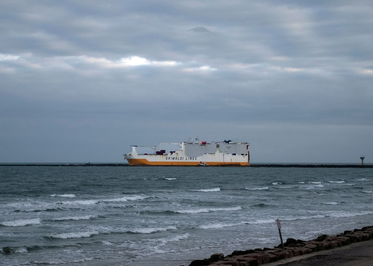 A large cargo ship sails across choppy waters under cloudy skies near a rocky coastline Freeport Surfside Texas