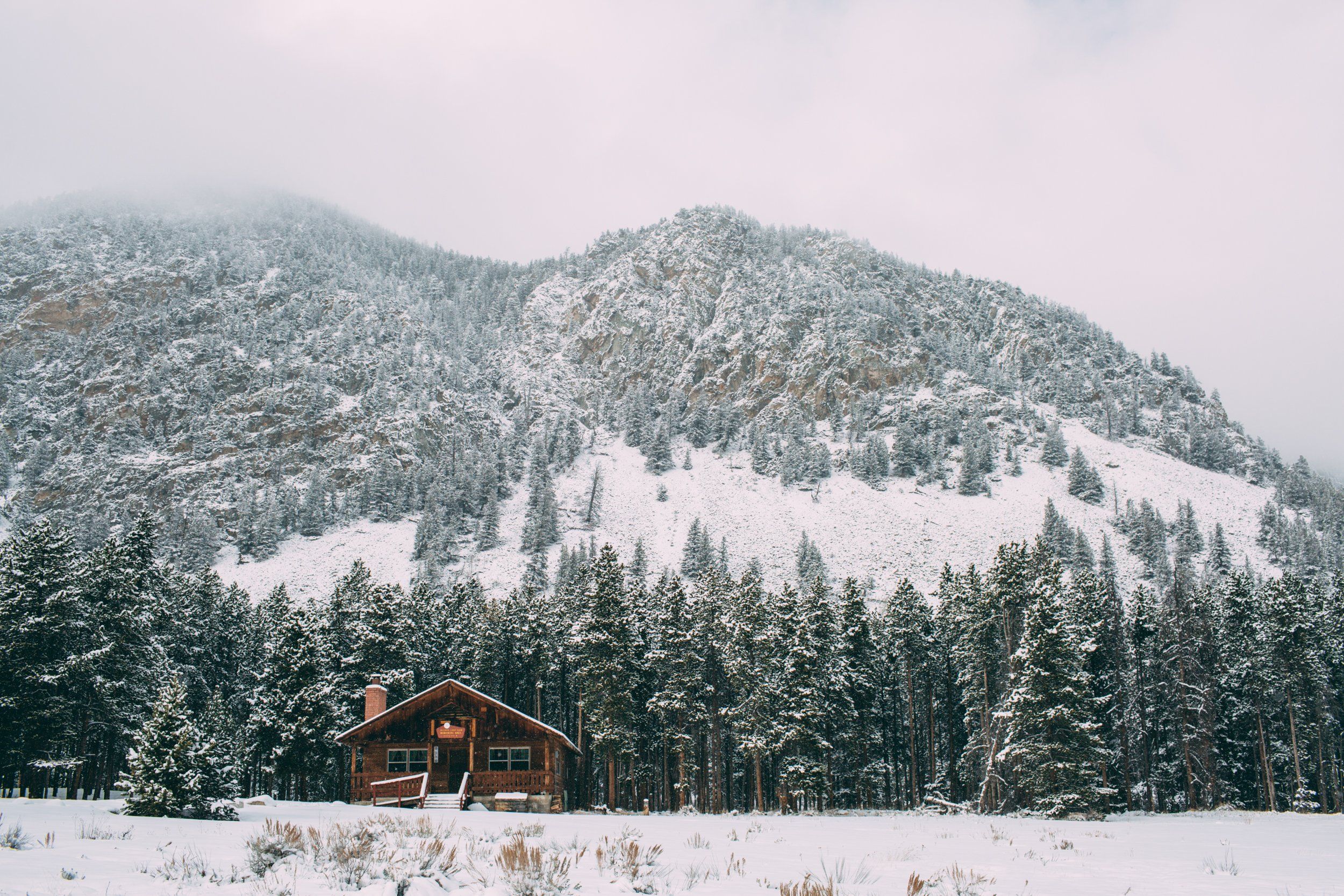 A snow-covered log cabin nestled among evergreen trees with snowy mountain peaks in the background.