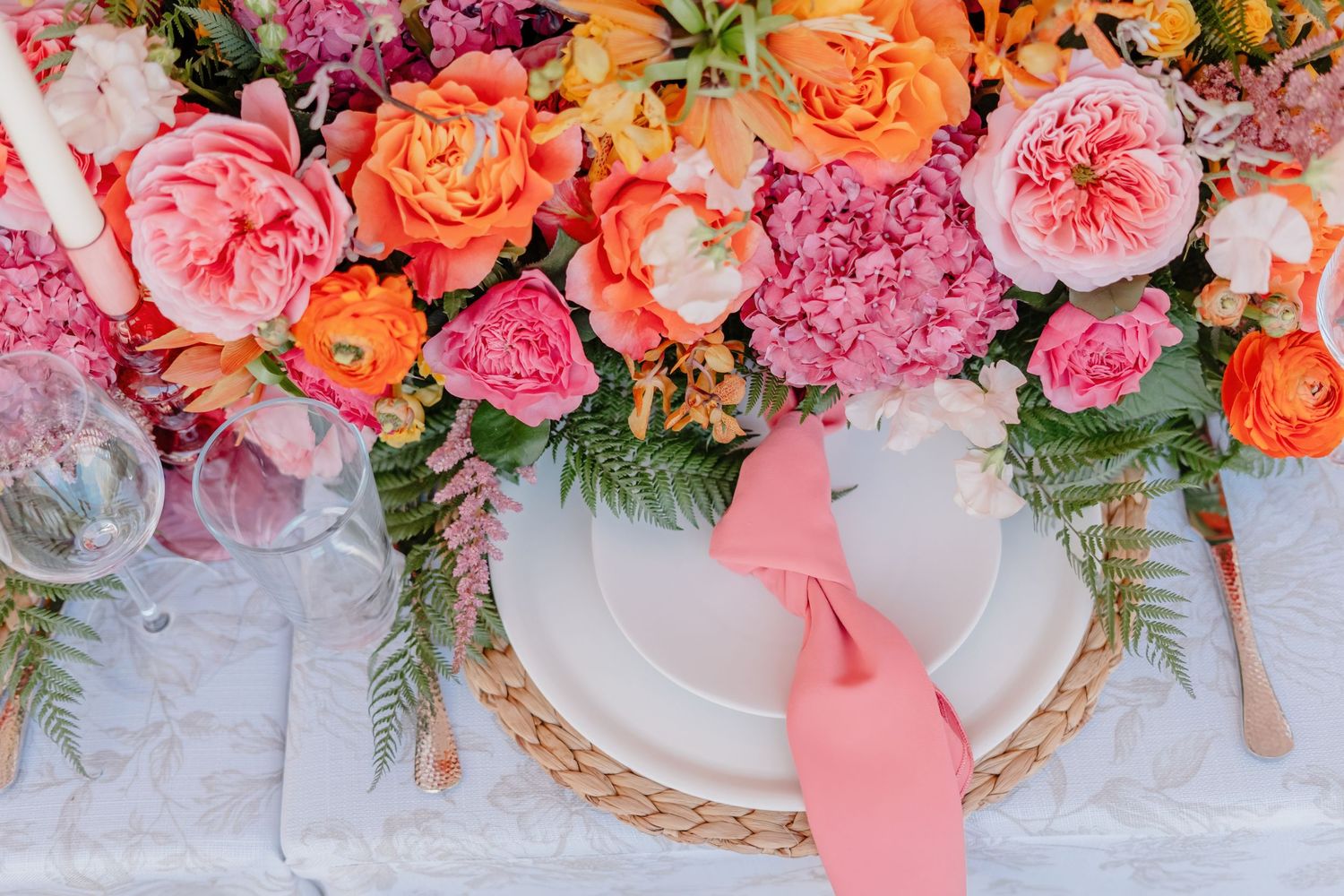 Overhead view of coral and pink roses with ferns decorating a place setting with woven charger plate and pink napkin.