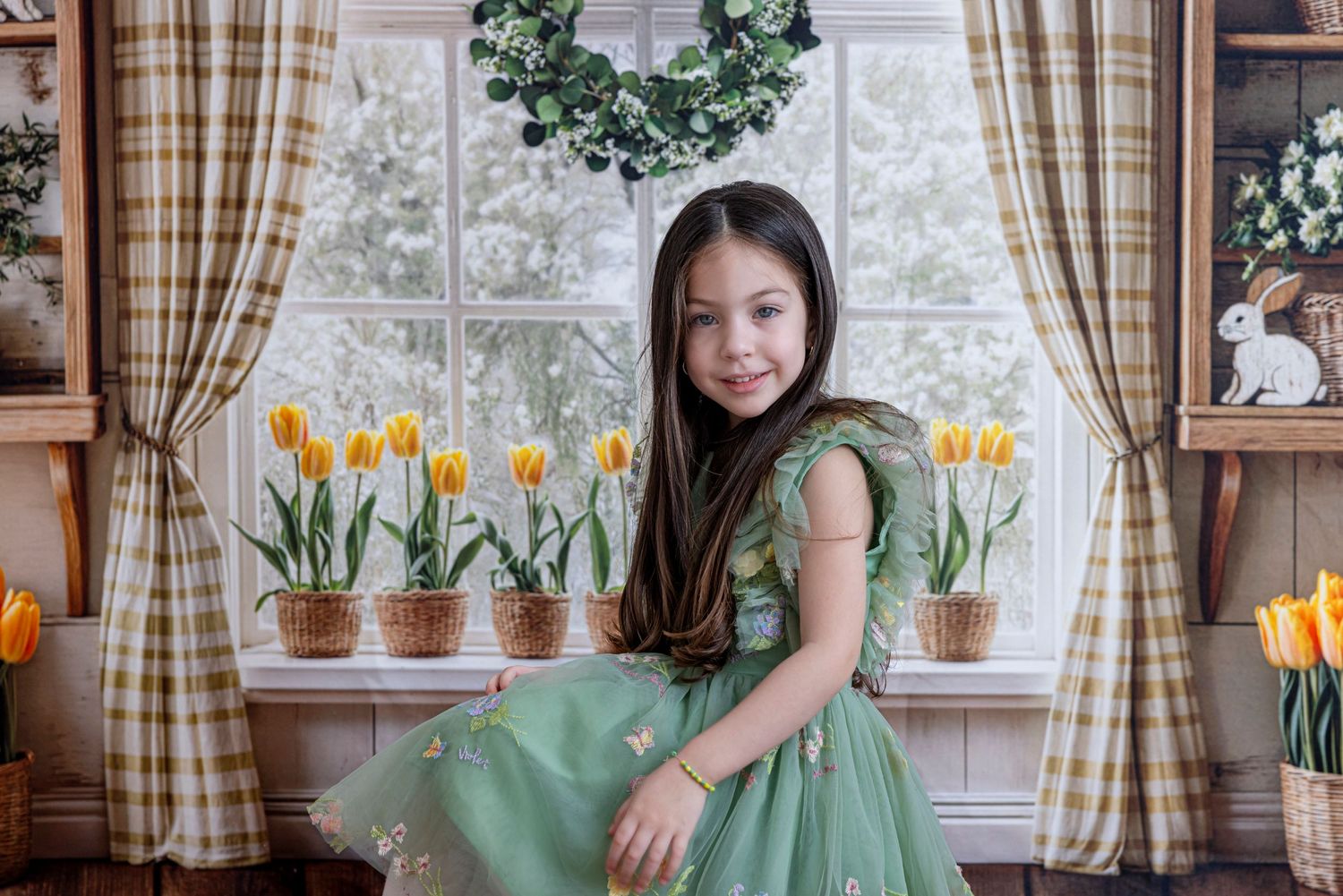 Child in green dress sits by window with potted yellow tulips and plaid curtains in cozy interior setting.