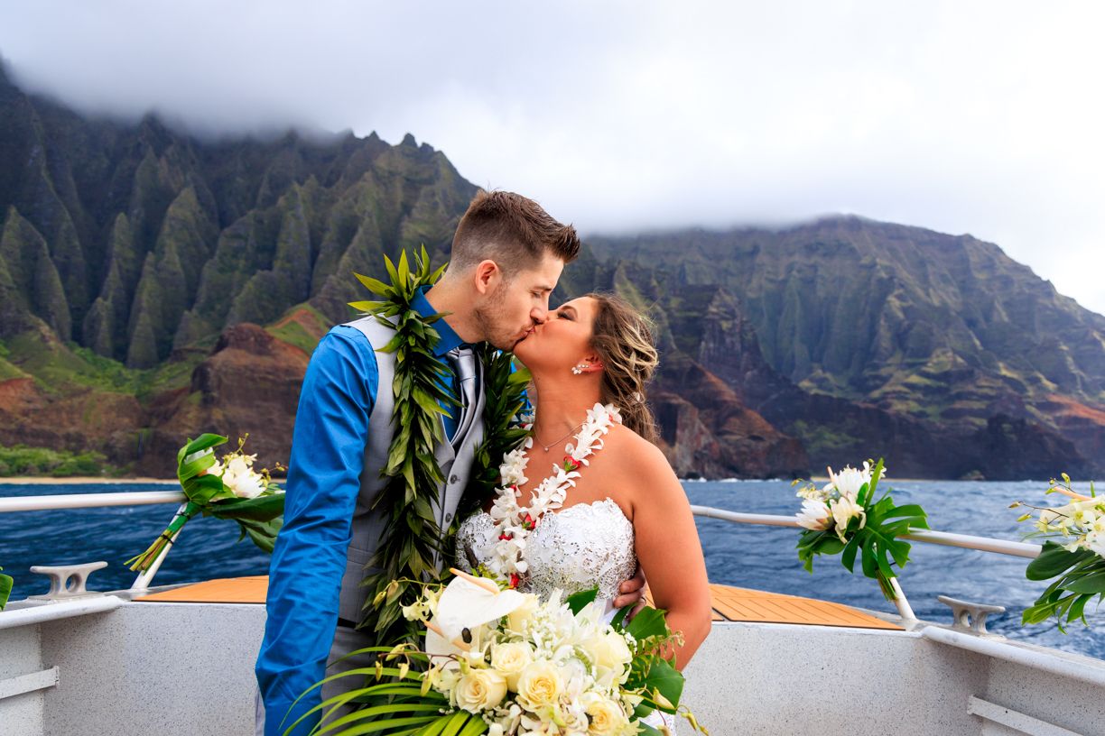 A romantic wedding couple shares a kiss on a boat deck with the stunning Na Pali Coast cliffs of Kauai in the background.