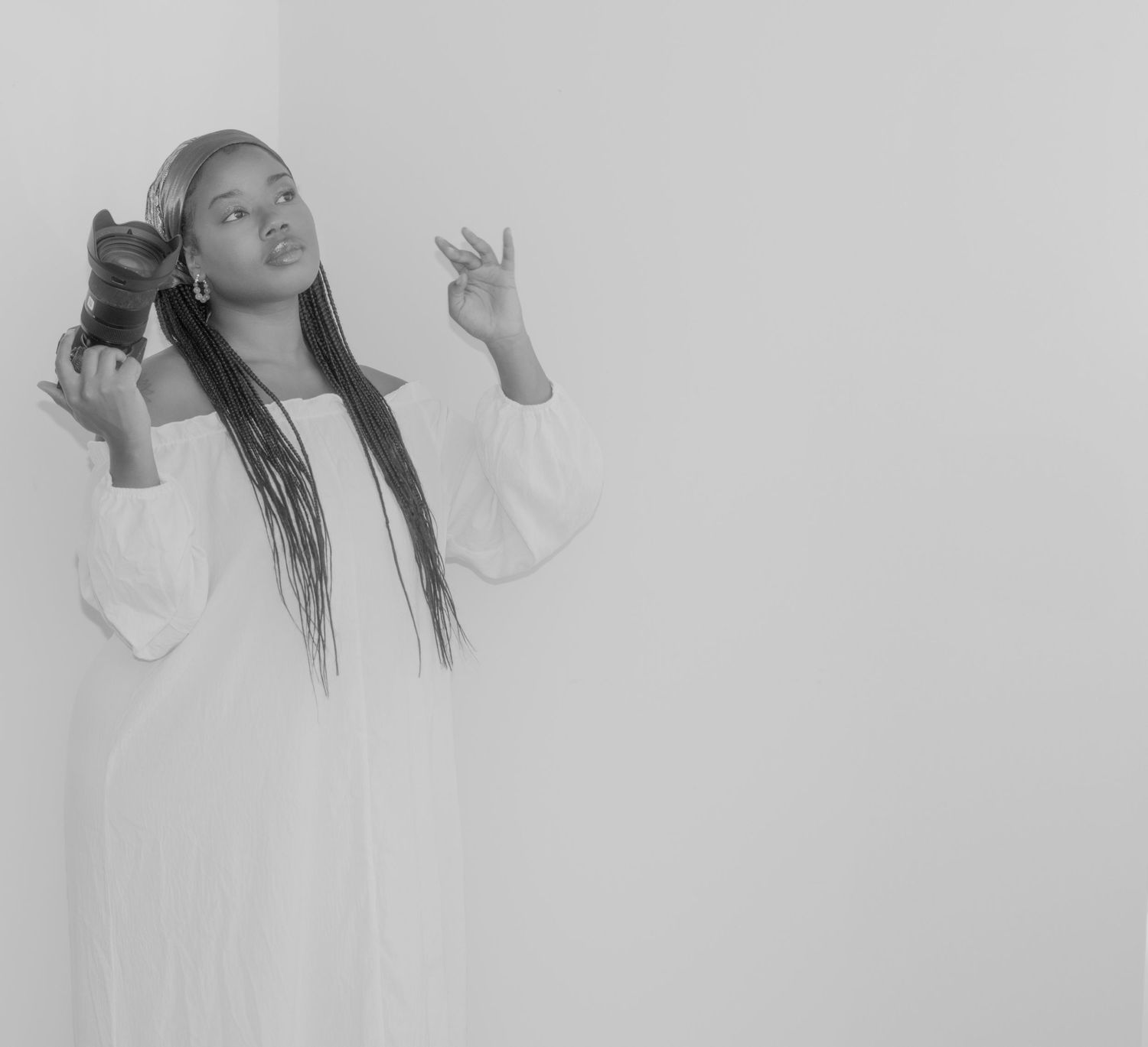 Black and white photo showing a person with long braids wearing a white dress making expressive gestures against a light background.
