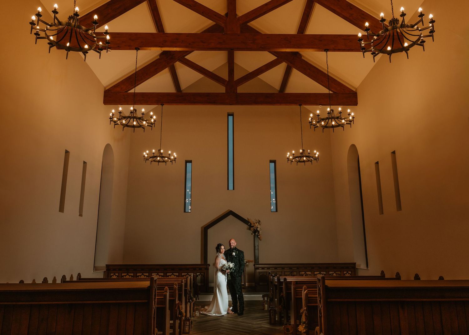 A couple stands silhouetted in a chapel with exposed wooden beams and hanging chandeliers creating a warm, romantic atmosphere.