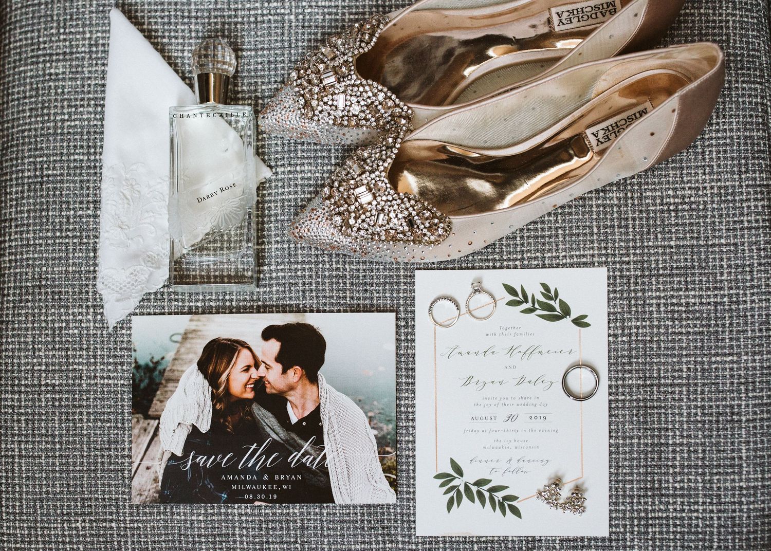 Wedding details including bridal shoes, rings, invitations and floral accessories arranged on textured gray fabric.