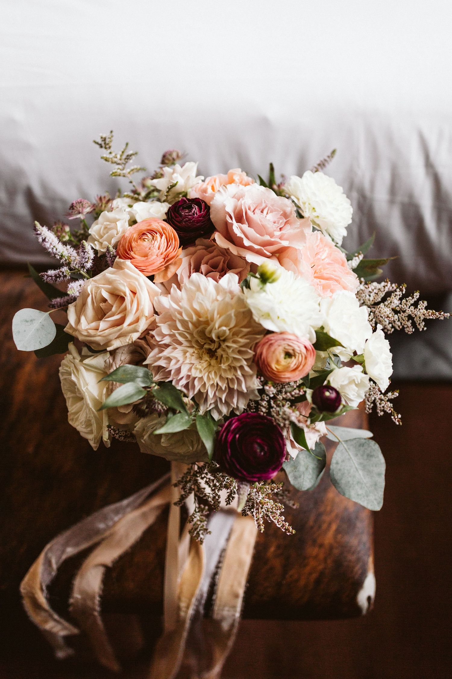 Romantic floral arrangement with peach roses, white flowers, and burgundy blooms tied with brown ribbon on dark surface.