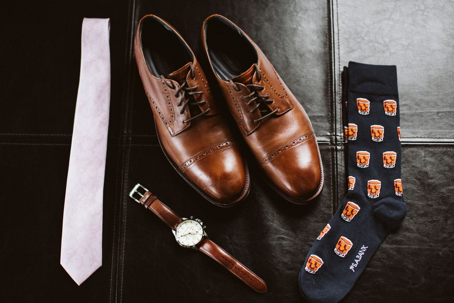 Wedding accessories including brown leather dress shoes, pink tie, patterned socks and wristwatch on dark surface.