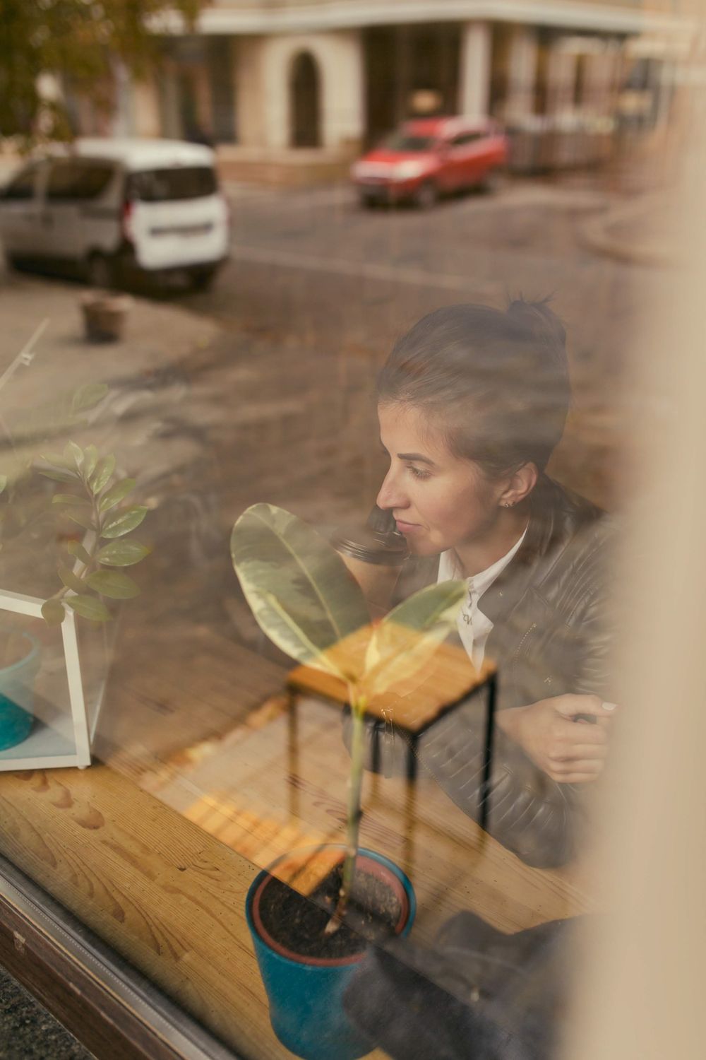 View through cafe window of a person sitting at wooden table with potted plant and warm lighting.