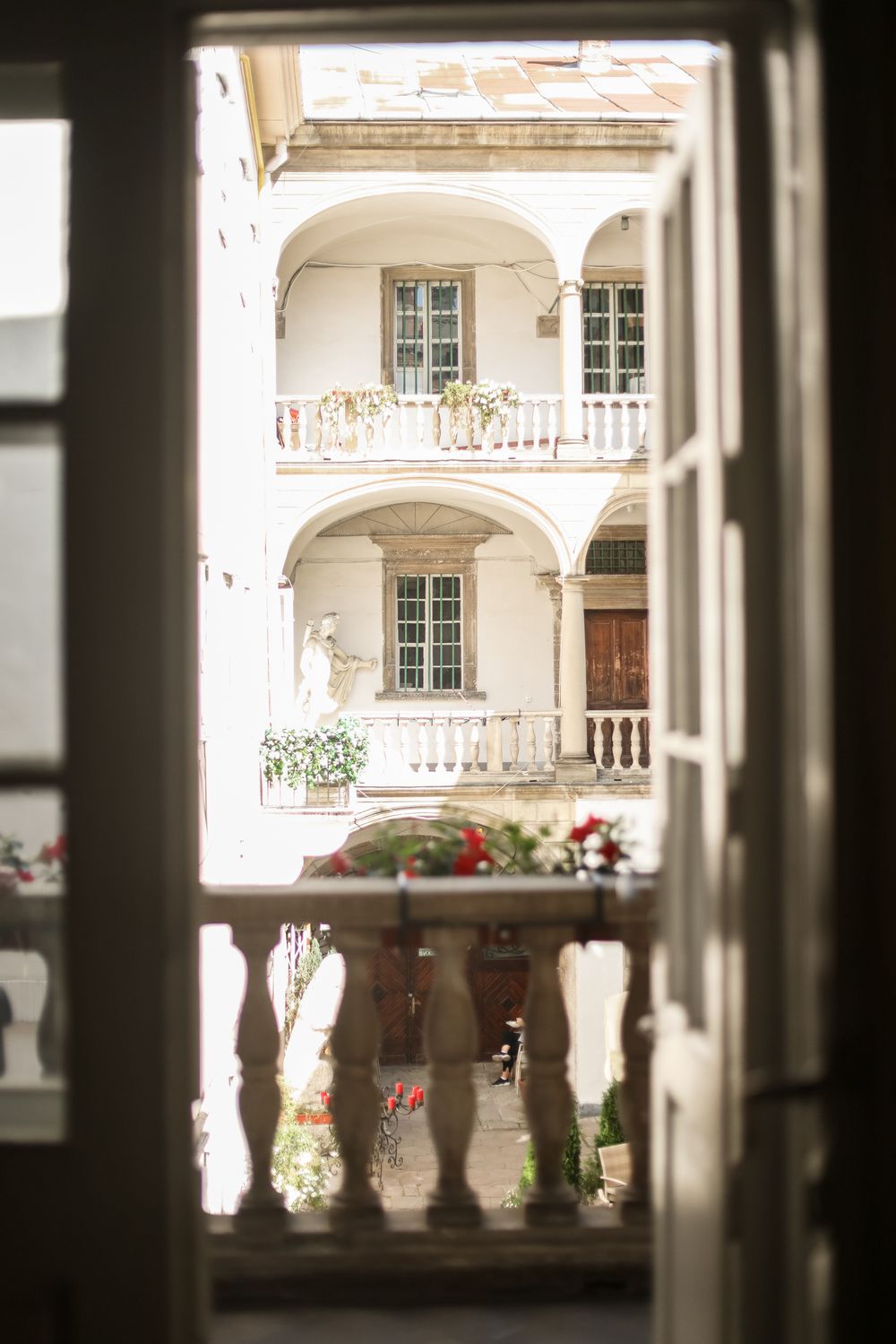 View through arched windows of an elegant white courtyard with classical architecture and red flowers on balustrades.