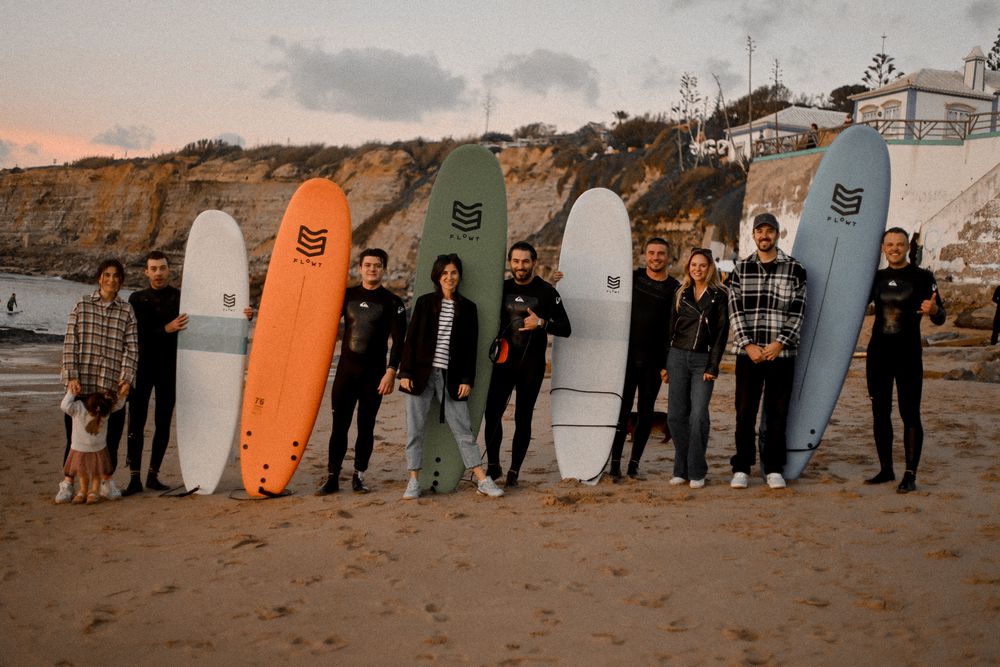 A group of surfers stand together on a sandy beach at sunset holding colorful surfboards against a cliffside backdrop.