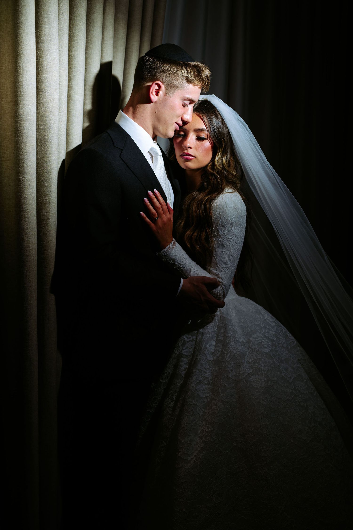 A bride and groom share a romantic moment in front of a dark curtain backdrop with dramatic lighting.