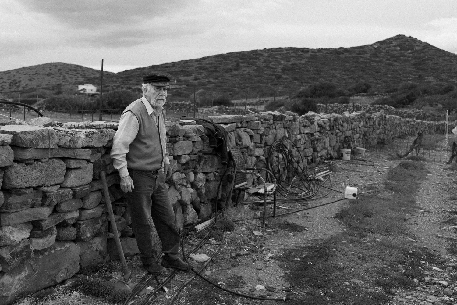 Black and white portrait of elderly farmer standing by stone wall in rural landscape.