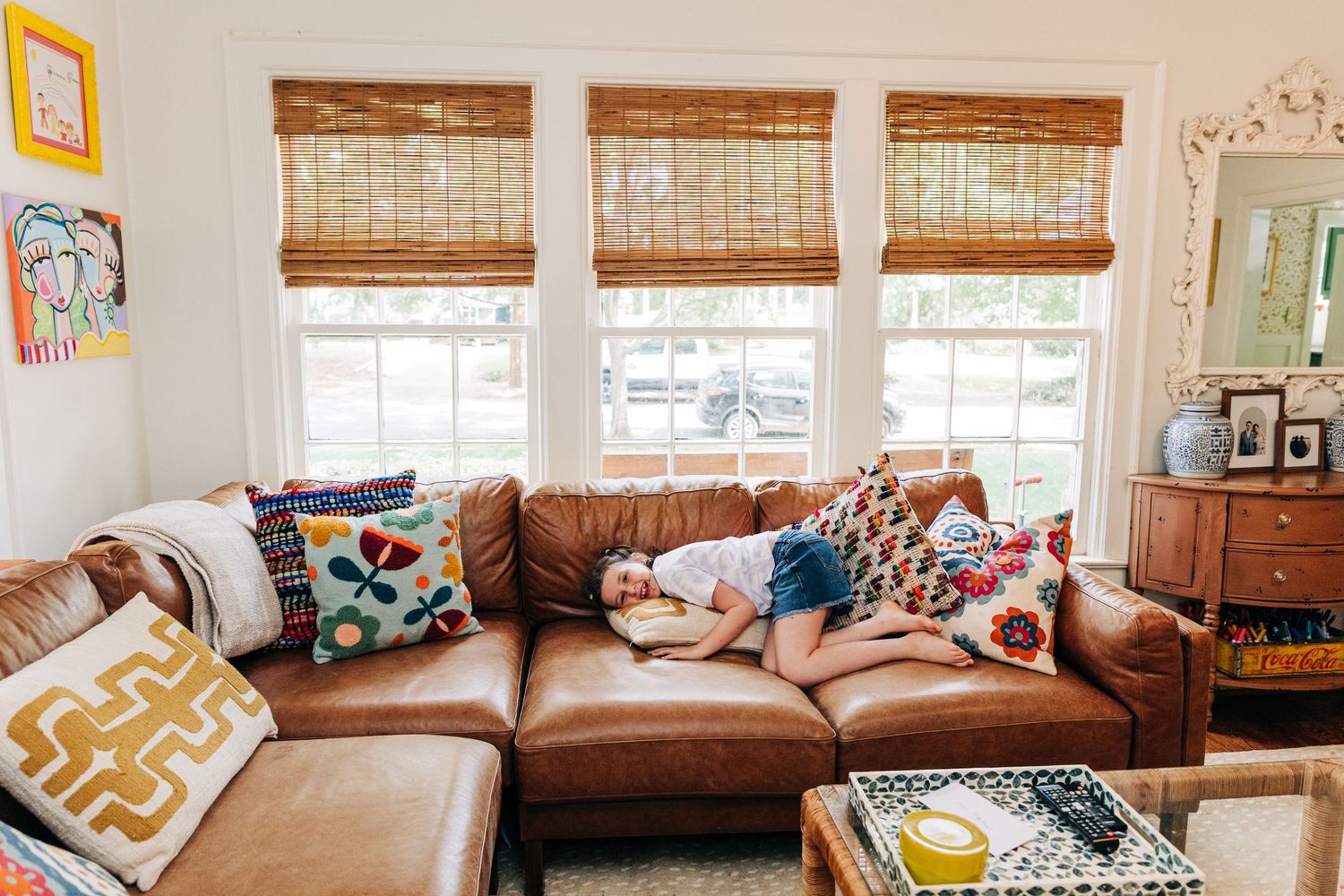 Cozy living room with brown leather sectional sofa, bamboo window shades, and colorful throw pillows by natural light, a young girl lays on.