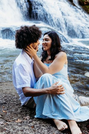 A romantic couple sitting together near a waterfall, sharing a tender moment with the woman holding the mans face.