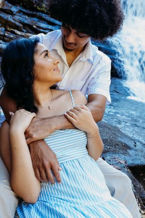 A loving couple cuddling near a waterfall, the man wrapping his arms around the woman in a blue striped dress.