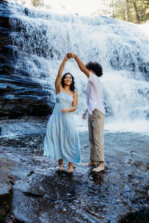 A couple dancing barefoot in front of a stunning waterfall, the man twirling the woman in a flowing blue dress.