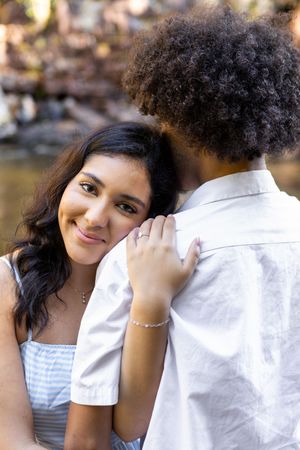 A smiling woman in a blue dress embraces her partner outdoors, looking warmly at the camera in natural light.
