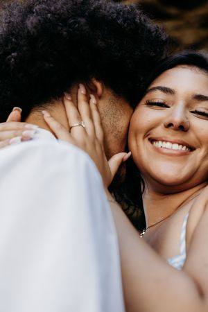 A woman with a bright smile embraces her partner closely, wearing a ring and necklace in a warm outdoor setting.