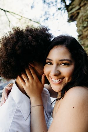 A beaming woman in a blue dress hugs her curly-haired partner outdoors, smiling joyfully at the camera.